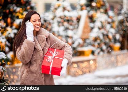 Beautidul girl with present near Christmas tree talks by phone in the snow outdoors. Happy girl near fir-tree branch in snow for new year.