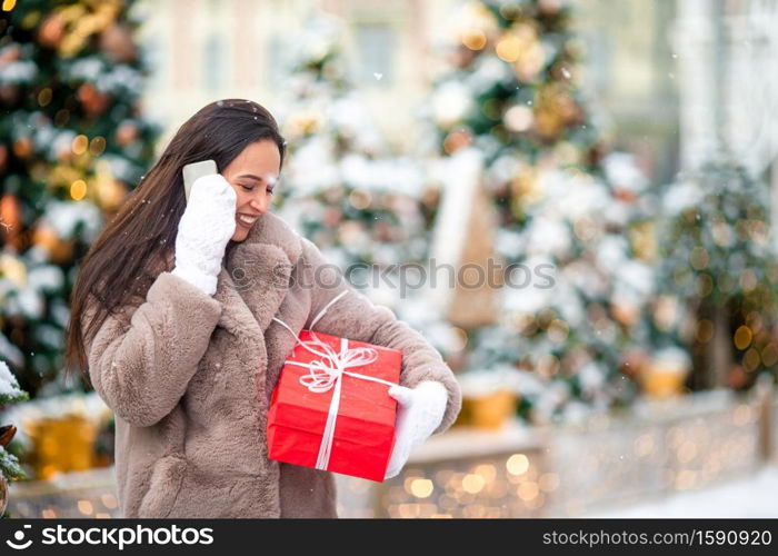 Beautidul woman near Christmas tree in the snow talking by phone with big red present. Happy girl near fir-tree branch in snow for new year.