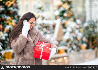 Beautidul woman near Christmas tree in the snow talking by phone with big red present. Happy girl near fir-tree branch in snow for new year.