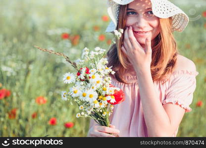 Beautieful young girl in the field of wild flowers. Teenage girl picking the spring flowers in the meadow, holding bouquet of flowers. She wearing hat and summer clothes. Spending time close to nature