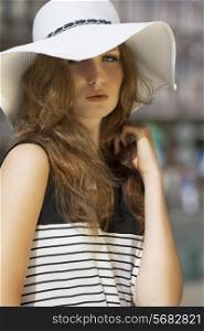 Beautifil, elegant woman in summer hat. She is on street in striped dress. She has brown makeup, and curly hair.