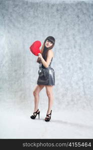 Beautiful 20-25 years young brunette woman holding red heart pillow on grey background