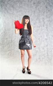 Beautiful 20-25 years young brunette woman holding red heart pillow on grey background