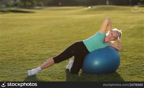 Beautiful active adult blonde woman doing side crunches on exercise ball, training abdominal muscles on park lawn at sunset. Active blonde lady in sportswear working out outdoors, exercising her abs on blue fitball.