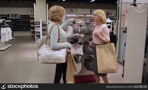 Beautiful adult blonde female friends with shopping bagschoosing brassiere in underwear department in clothing store. Joyful senior women buying sexy lace lingerie in boutique shop during shopping together. Slow motion.