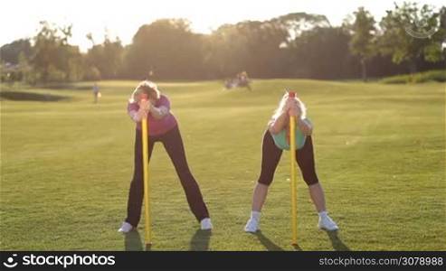 Beautiful adult fitness ladies in sports clothes doing exercises with body bars in the park. Pretty sporty senior females training with fitbars outdoors, stretching and bending upward in rays of setting sun.