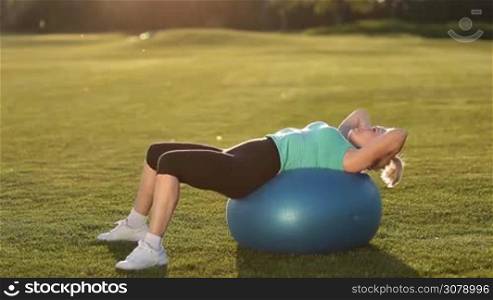 Beautiful adult sportswoman doing abdominal crunches on fit ball while training on park lawn at sunset. Smiling senior fit female in sportswear exercising abs with exercise ball. Side view.