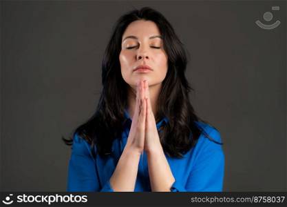 Beautiful adult woman praying over grey background. Lady in blue shirt begging someone with hope in eyes. Beautiful adult woman praying over grey background. Lady in blue shirt begging someone with hope in eyes.