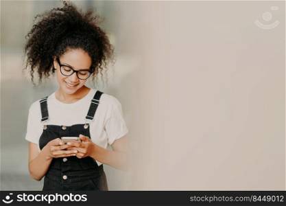 Beautiful African American girl in fashionable clothing, uses mobile phone for sending text messages, connected to wifi, checks email box, stands near white wall with empty space. Online communication