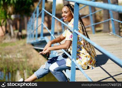 Beautiful African girl combed with colorful braids sitting on an urban bridge with a camera on vacation.. African girl combed with braids sitting on an urban bridge with a camera on vacation