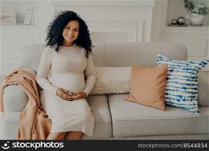 Beautiful african pregnant young woman sitting on sofa decorated with cushions, feeling happiness and love, keeping hands crossed on her bump belly in living room ambience, relaxing at home. Beautiful african pregnant young woman feeling happiness and love while spending time at home