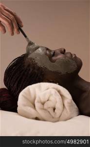 Beautiful african woman having clay facial mask applying by beautician