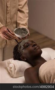 Beautiful african woman having clay facial mask applying by beautician