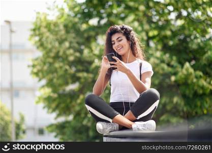 Beautiful African woman listening to music with earphones and smart phone outdoors. Arab girl in sport clothes with curly hairstyle in urban background.