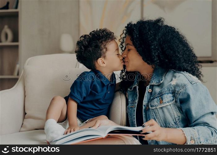 Beautiful afro american family mother with little son kissing each other and expressing love while spending time together at home, reading book while sitting on soft white couch in living room. Happy mixed race young mother with little child kissing each other while spending time together at home