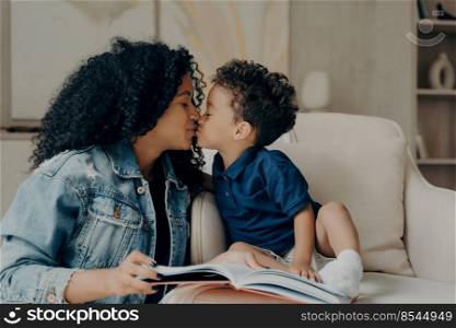 Beautiful afro american family mother with little son kissing each other and expressing love while spending time together at home, reading book while sitting on soft white couch in living room. Happy mixed race young mother with little child kissing each other while spending time together at home