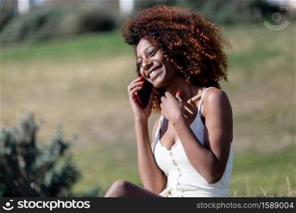 Beautiful afro american woman with white dress sitting on grass in a park in sunny day while using smartphone