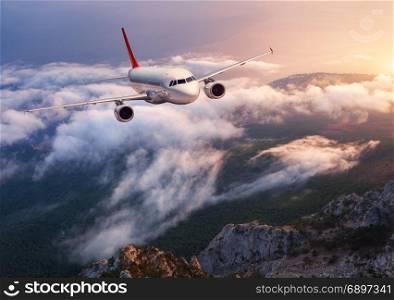 Beautiful airplane is flying over low clouds at sunset. Landscape with passenger airplane, rocks, forest, blue sky with orange sunlight in dusk. Passenger aircraft. Business travel. Commercial plane