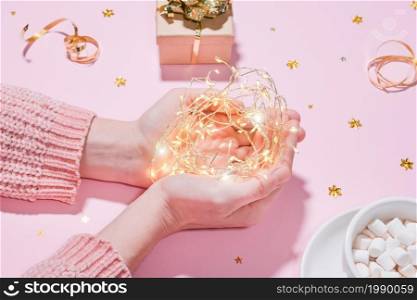 Beautiful and cozy holiday concept Hands holding string garland lights, on the background of wrapped gift, a cup of hot chocolate or cocoa with marshmallows, golden tinsel isolated on pink background. Beautiful and cozy holiday concept. Hands holding string garland lights, on the background of wrapped gift, a cup of hot chocolate or cocoa with marshmallows, golden tinsel isolated on pink background