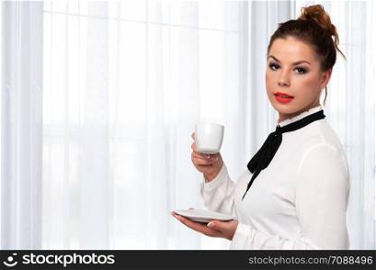 Beautiful and elegant woman holding a cup of coffee in her hands and enjoying its flavor on a background of luxury interior (copy space).