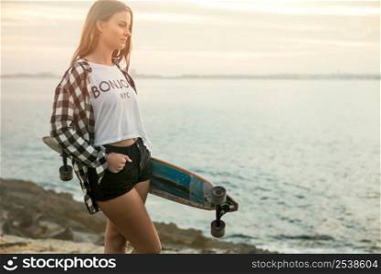 Beautiful and fashion young woman posing at the sunset with a skateboard