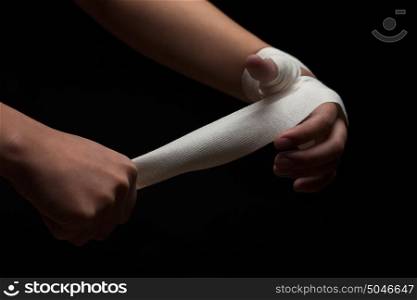 Beautiful and fit female fighter getting prepared for the fight or training, wrapping her hands with bandage tape against dark background.