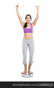Beautiful and happy athletic woman over a scale with arms up, isolated on white background. Healthy girl