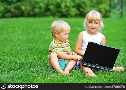 beautiful and happy little girl with brother sitting on grass with laptop computer