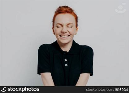 Beautiful and happy pleased red haired woman with closed eyes and toothy smile posing against grey wall. Cheerful excited female demonstrating positive emotions and happiness, dressed casually. Happy pleased red haired woman with closed eyes and toothy smile posing against grey wall
