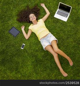 Beautiful and happy young woman lying on the grass surrounded by technology devices