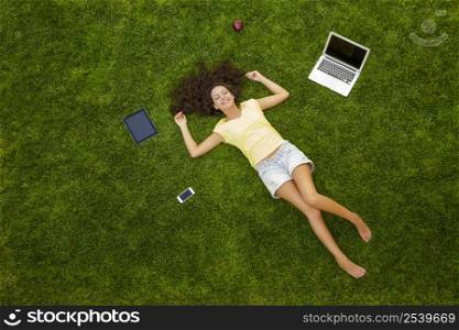 Beautiful and happy young woman lying on the grass surrounded by technology devices