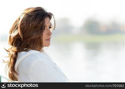 Beautiful and sensual woman is looking thoughtful at the beautiful landscape of the coast or river in the city illuminated by deep morning sunlight.