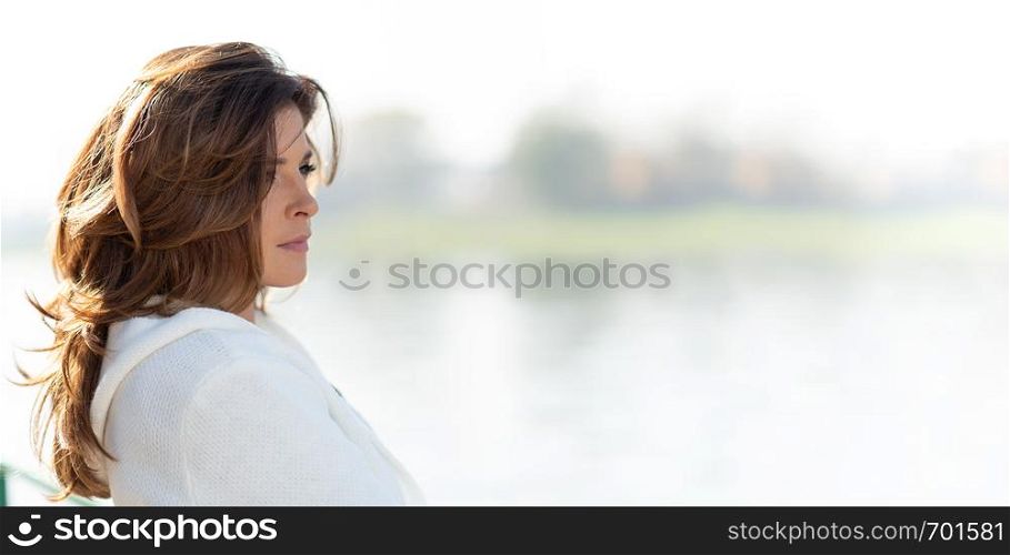 Beautiful and sensual woman is looking thoughtful at the idyllic landscape of the coast or river in the city illuminated by deep morning light (copy space)