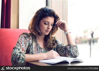 Beautiful and sensual young woman reading a book next to a window in a restaurant or coffee bar.