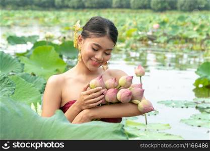 Beautiful asia women wearing traditional Thai dress and sitting on wooden boat in flower lotus lake. Her hands are holding a pink lotus and picking up flower. And her face is be happy and smile.
