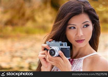 Beautiful Asian Chinese girl or young woman wearing summer dress outside in green environment taking pictures or photographs in Autumn or Fall with a retro style camera