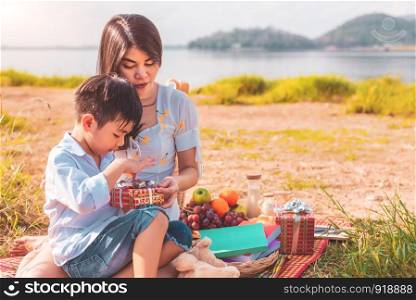 Beautiful Asian mother and son doing picnic and opening gift box from surprise in Birthday party on meadow near lake and mountain. Holiday and Vacation. People lifestyle and Happy family life concept