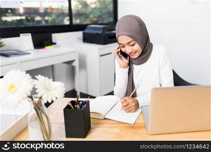 Beautiful Asian muslim woman working using laptop and mobile phone call in modern office. Small business company owner, startup entrepreneur, or working woman lifestyle concept
