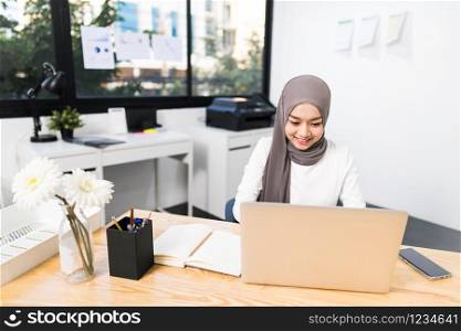 Beautiful Asian muslim woman working using laptop in modern office. Small business company owner, startup entrepreneur, or working woman lifestyle concept