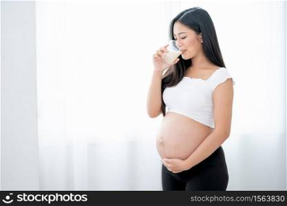 Beautiful Asian pregnant woman drink milk and stand in front of white curtain and she look happy with morning light. Concept of good healthy food for mother and support baby in belly of people.
