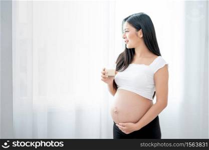 Beautiful Asian pregnant woman hold glass of milk and look outside through white curtain and she look happy by smiling. Concept of good healthy food for mother and support baby in belly people.