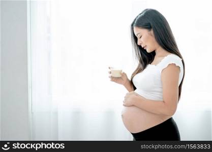 Beautiful Asian pregnant woman hold glass of milk and look to her belly in front white curtain and she look happy by smiling. Concept of good healthy food for mother and support baby in belly people.