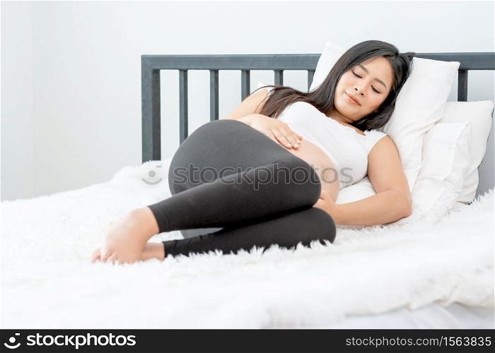 Beautiful Asian pregnant woman lie on bed and look relax with morning light in bedroom. Concept of good healthy activity for mother and support growth of baby in belly of people.