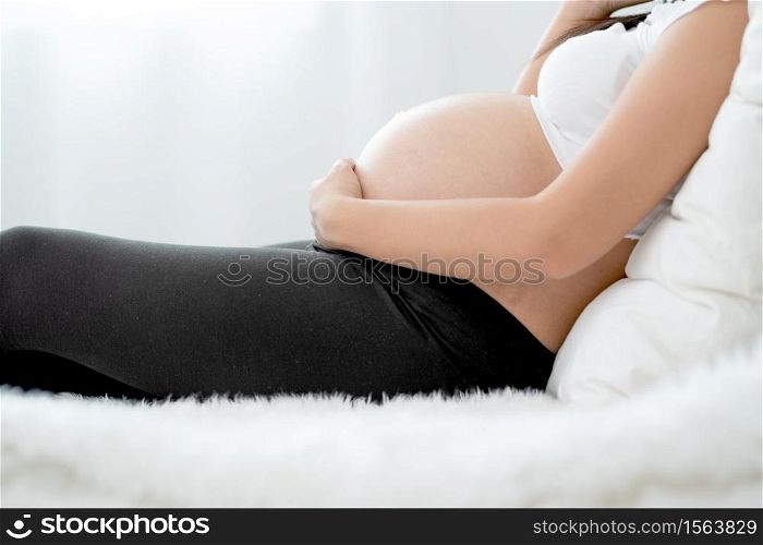 Beautiful Asian pregnant woman lie on bed and use hand touch belly with carefully emotion. Concept of good healthy activity for mother and support growth of baby in womb of people.