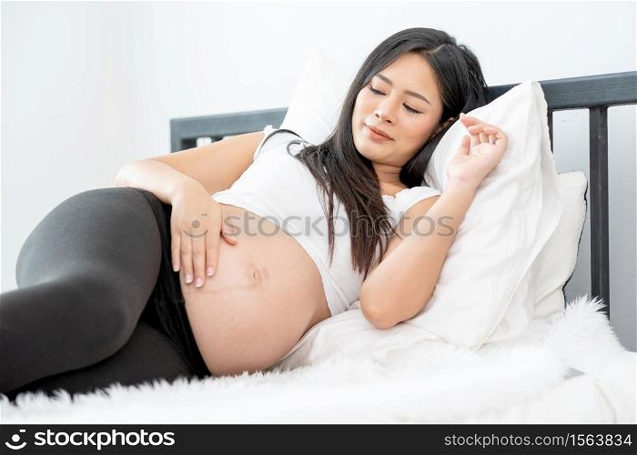 Beautiful Asian pregnant woman lie with relax position and use one hand touch her belly. Concept of good healthy activity for mother and support growth of baby in womb of people.