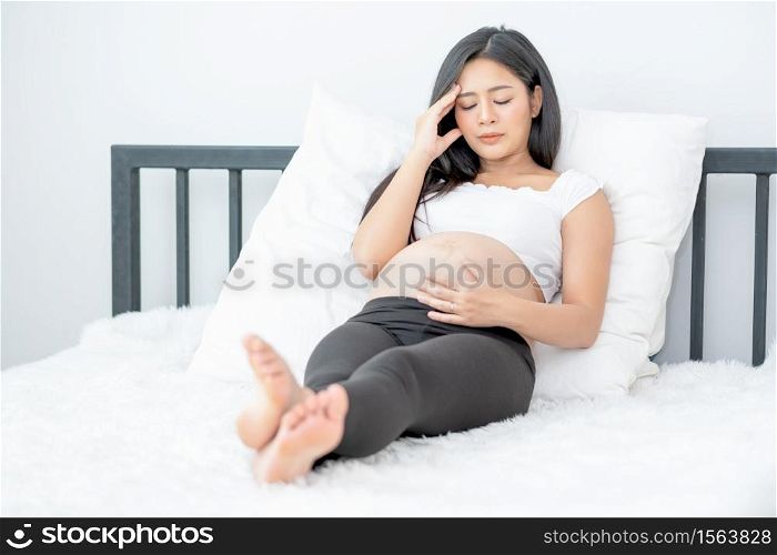 Beautiful Asian pregnant woman show action of stress and headache lie on bed with morning light. Concept of sickness of mother during pregnancy and need to take care and support at home.