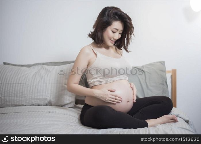 Beautiful asian pregnant woman sit on bed and touching her belly