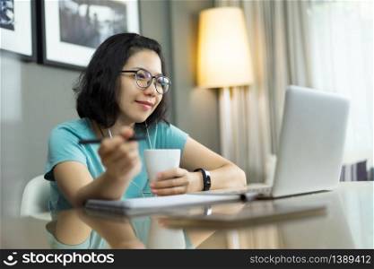 Beautiful Asian professional woman with blue shirt and eyeglasses in confident expression while work from home. Attractive young girl working on laptop computer during quarantine and isolated. Prevention Pandemic virus. Covid 19, Coronavirus, Healthy.