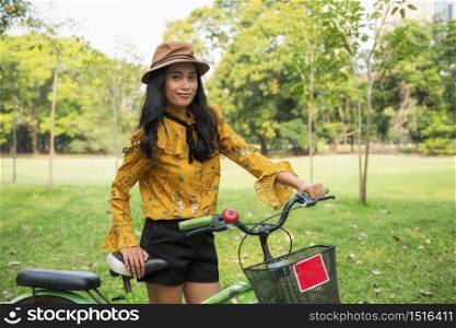 Beautiful Asian tan Bohemian woman with yellow vintage dress and hat hold bicycle to ride bike in outdoor park. Summer activity for attractive healthy girl with Boho costume with greenery background.
