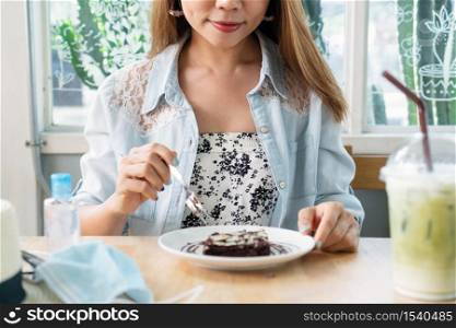 Beautiful Asian woman eating brownie cake with iced Matcha latte, surgical mask, hand sanitize on wooden table at cafe in sunny day. Close up.